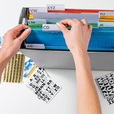 Hände sortieren farbige Akten in einem Ordner mit beschrifteten Trennblättern. Daneben liegen Aufkleber mit Buchstaben.