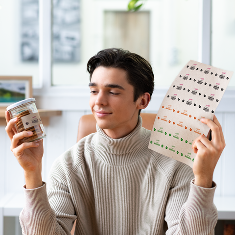 Ein junger Mann hält in der einen Hand ein Glas mit Snacks und in der anderen Hand ein Etikett mit verschiedenen Designs.