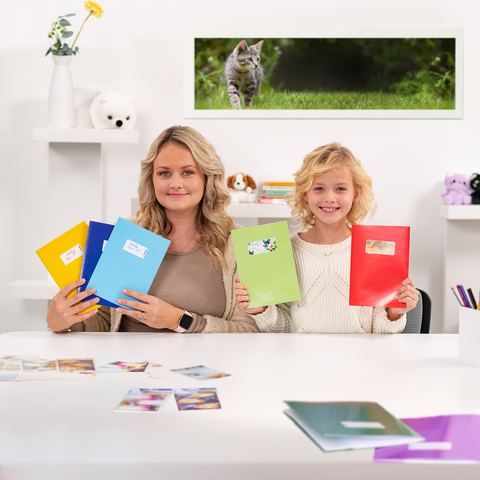 Eine Frau und ein Mädchen halten bunte Ordner in der Hand, während sie an einem Tisch mit Fotos sitzen. Im Hintergrund ist eine Katze zu sehen." 

(Note: The text is already in German and does not require translation. It is clear and fluent as it is.)