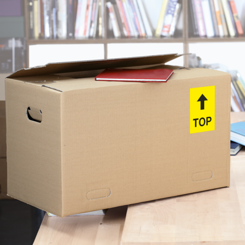 Kartonbox mit einem roten Heft auf der Oberseite, beschriftet mit einem gelben 'TOP'-Schild, steht auf einem Tisch in einer Bibliothek." 

(Note: The text is already in German and does not require any changes for tone, context, or meaning.)